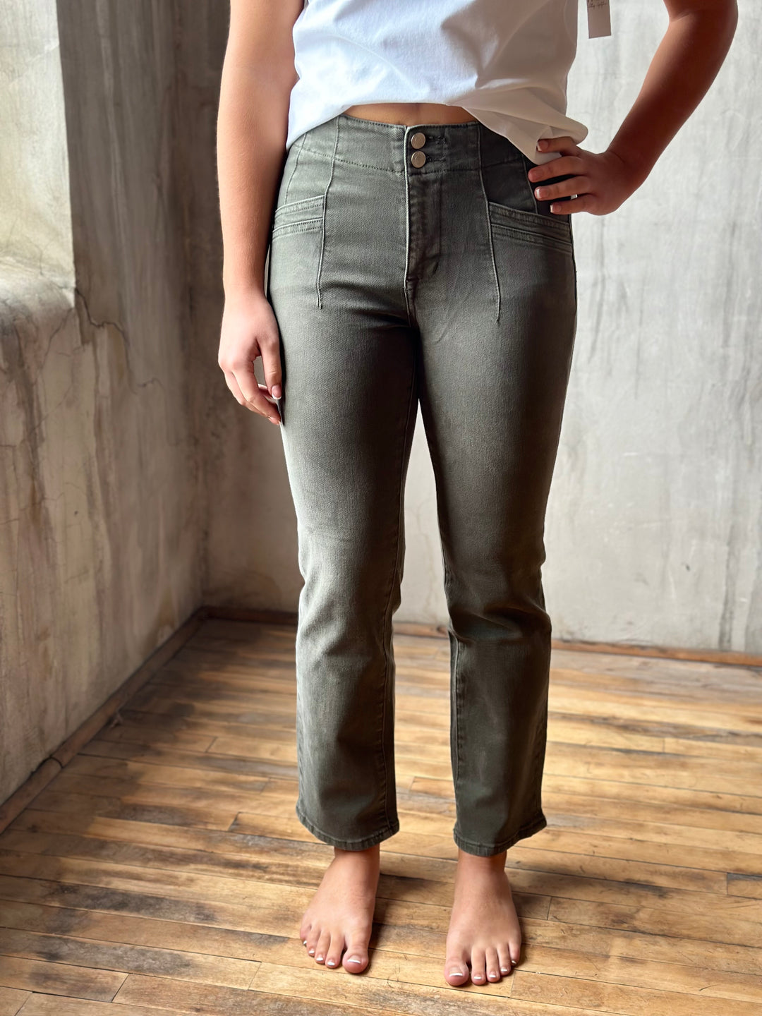 A person wearing olive green straight-leg jeans, standing barefoot on a wooden floor against a textured concrete wall.