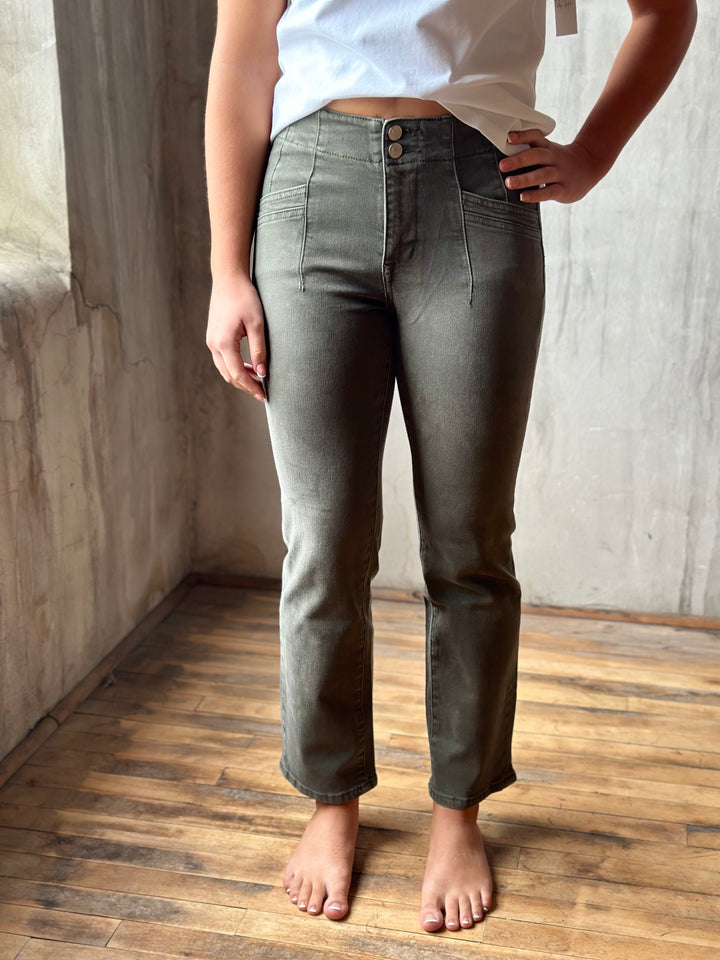 A person wearing olive green straight-leg jeans, standing barefoot on a wooden floor against a textured concrete wall.