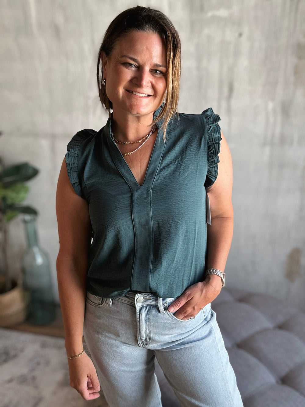 Woman wearing a dark green v-neck sleeveless top with ruffles on the sleeves and light blue jeans standing indoors.