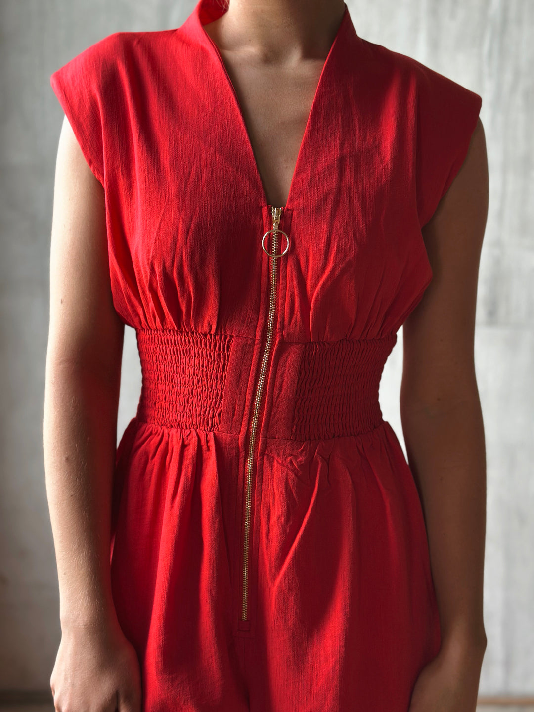 Close up Front view of red sleeveless jumpsuit features a flattering v-neckline, a front zipper closure, and a smocked waist