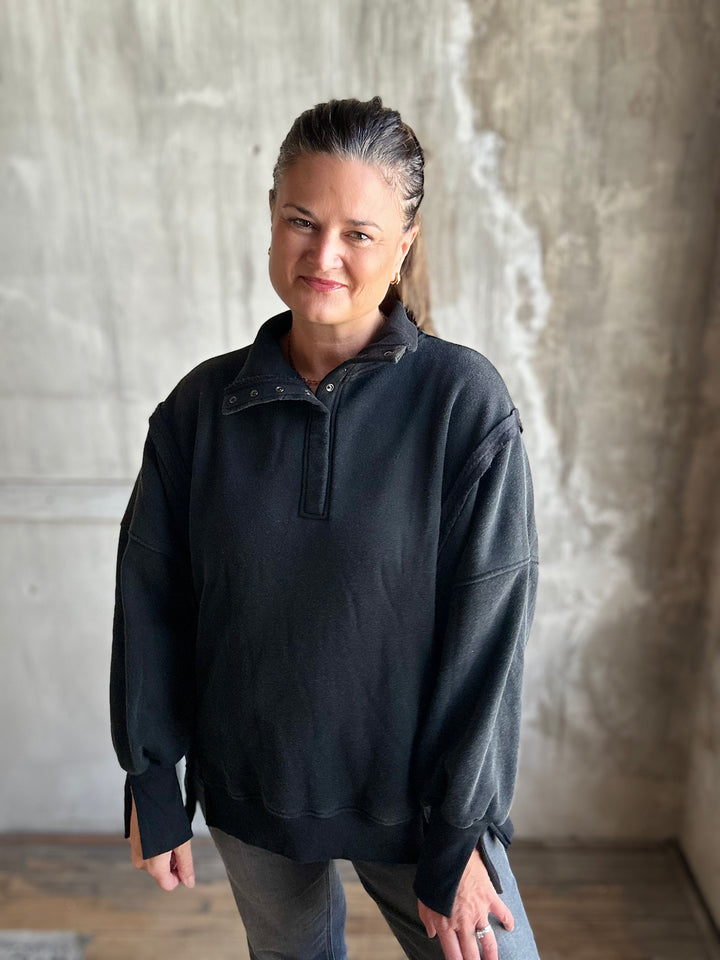 A woman wearing a faded black half-placket pullover sweatshirt and gray jeans, standing against a textured concrete wall.