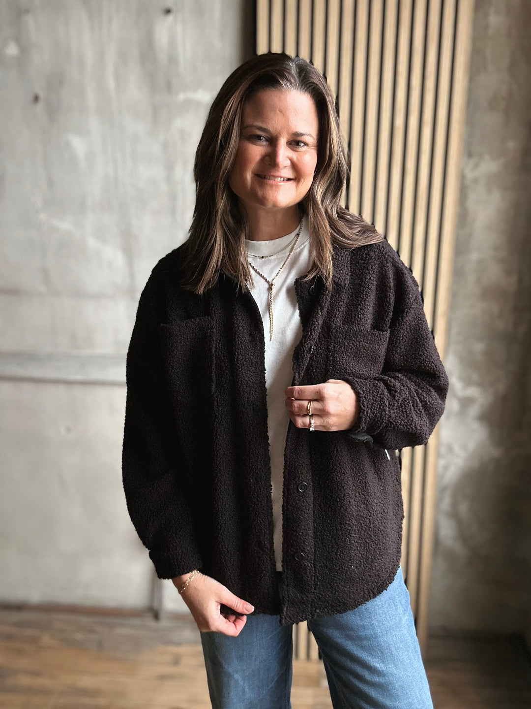 Woman wearing a black jacket and blue jeans standing against a concrete wall.