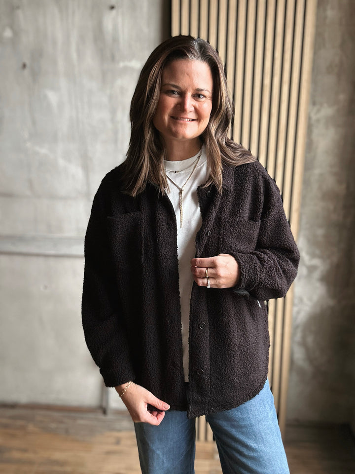 Woman wearing a black jacket and blue jeans standing against a concrete wall.