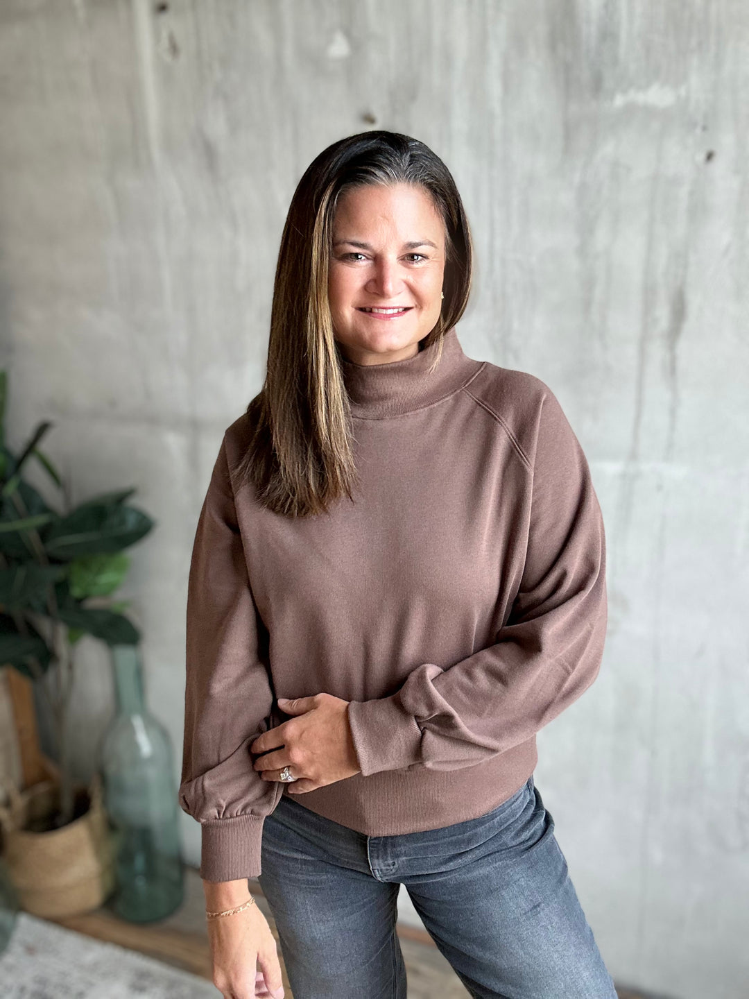 A woman with long brown hair wearing a taupe-brown mock neck sweatshirt and blue jeans, standing in front of a textured concrete wall with a plant in the background.