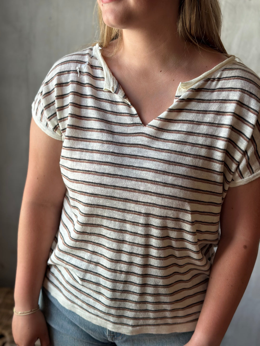 A person wearing a white and brown striped v-neck shirt with a neutral background.