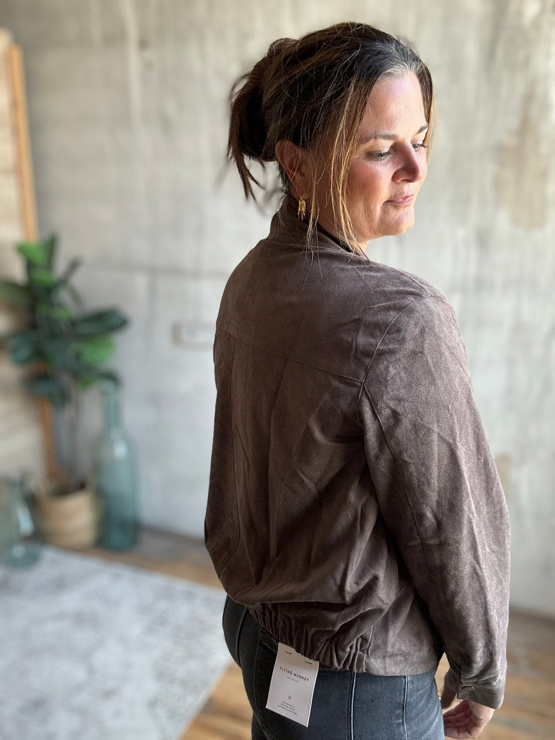 A woman wearing a brown textured jacket and blue jeans, photographed from the back in a rustic indoor setting with a concrete wall and plants in the background.