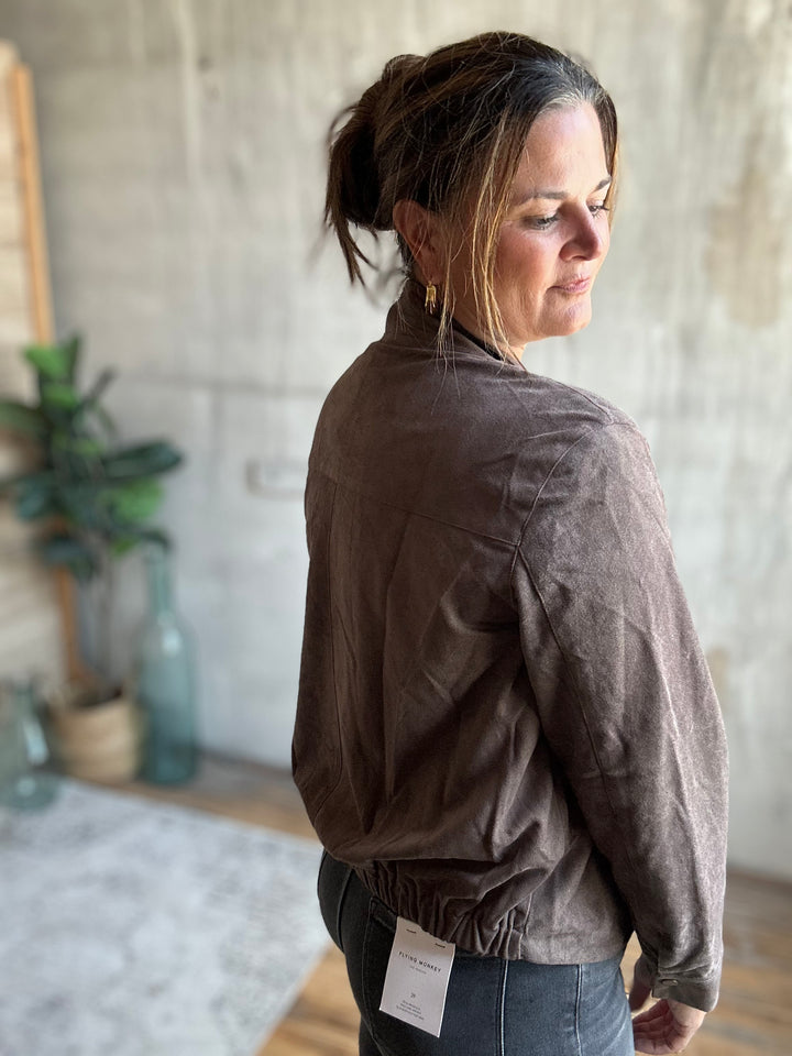 A woman wearing a brown textured jacket and blue jeans, photographed from the back in a rustic indoor setting with a concrete wall and plants in the background.