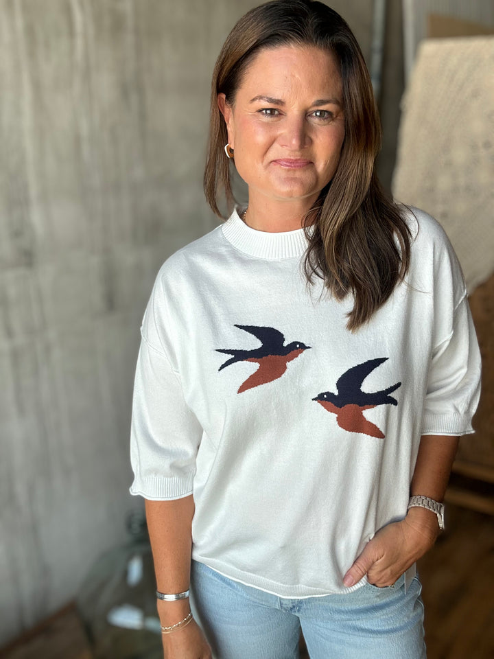 A woman wearing a white sweater with  swallow birds, paired with light blue jeans, standing in a softly lit indoor setting.