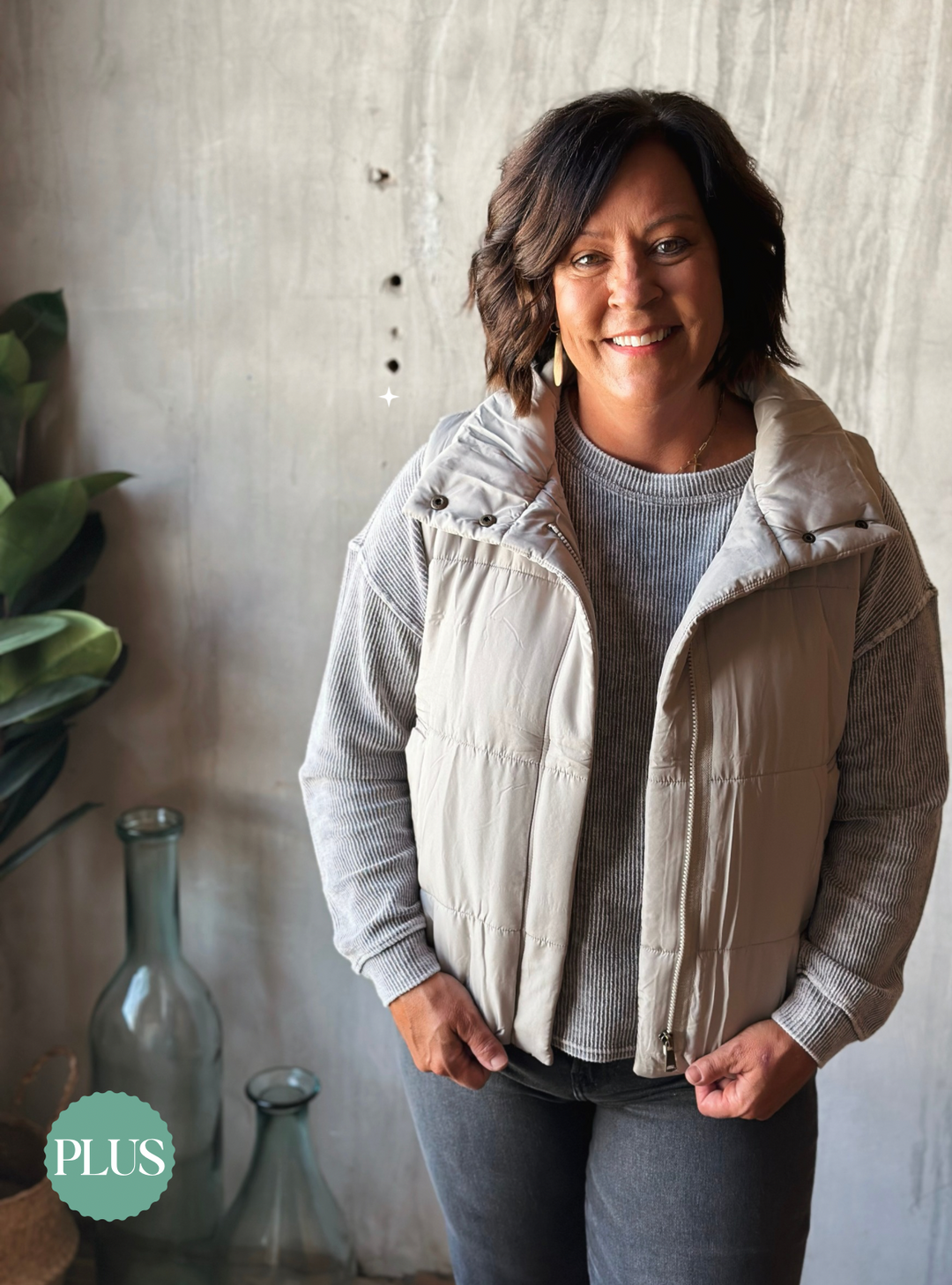 Woman wearing a beige vest over a gray sweater in a home setting with a plant and vase.