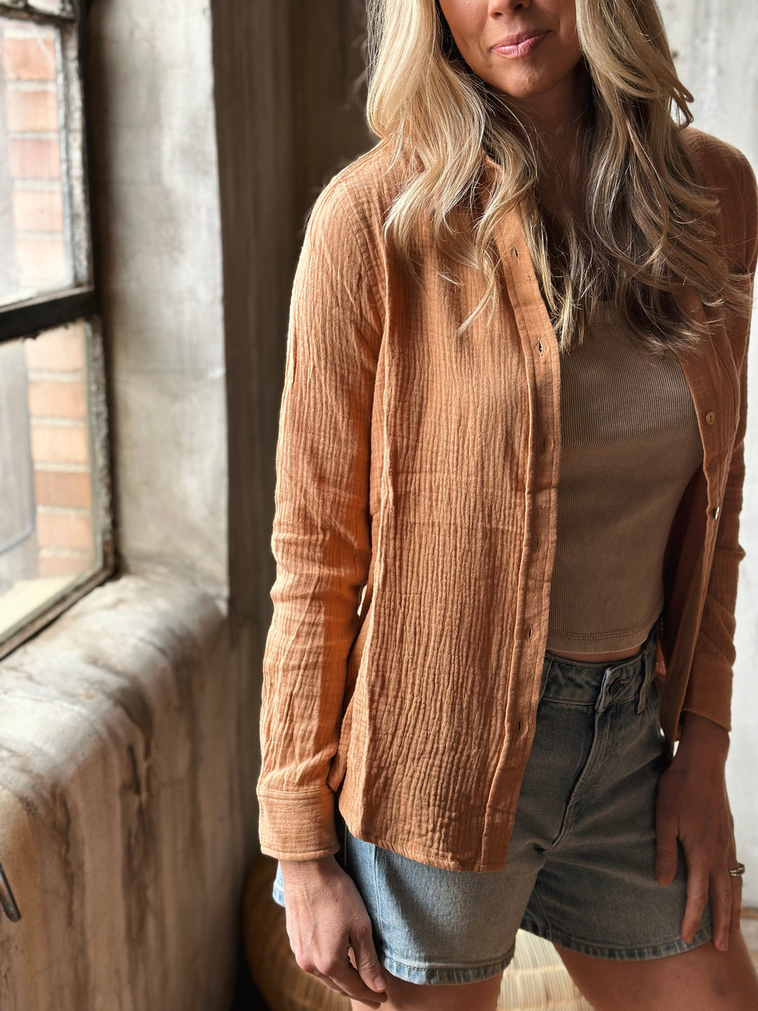 Woman wearing a burnt orange textured blouse and denim shorts standing by a window.