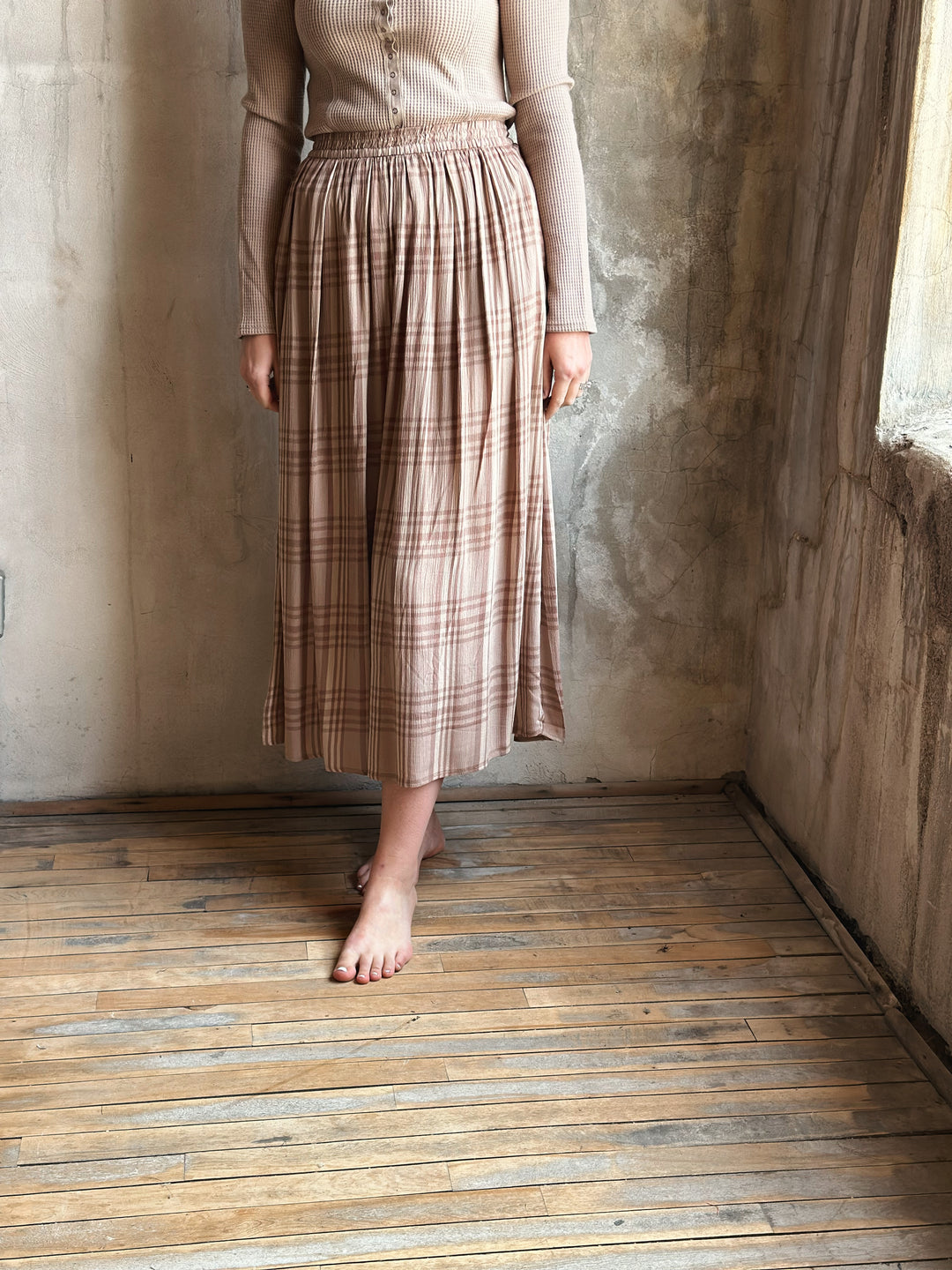 Person wearing a plaid skirt standing in a room with wooden floor and textured walls.