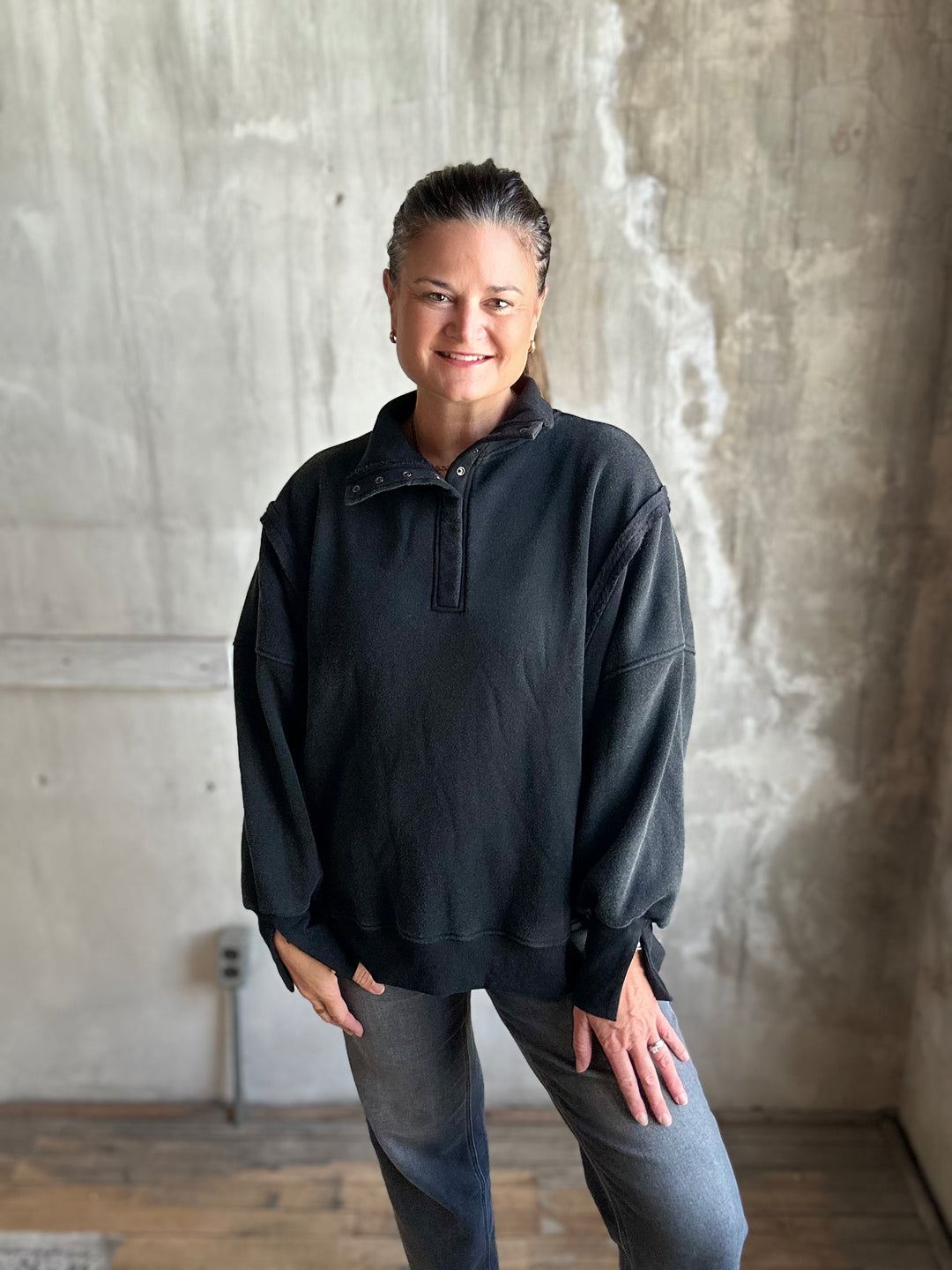 A woman wearing a black long-sleeve quarter-placket pullover and gray jeans, standing in front of a textured concrete wall.