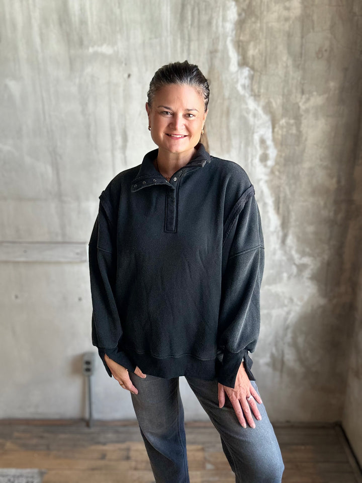 A woman wearing a black long-sleeve quarter-placket pullover and gray jeans, standing in front of a textured concrete wall.