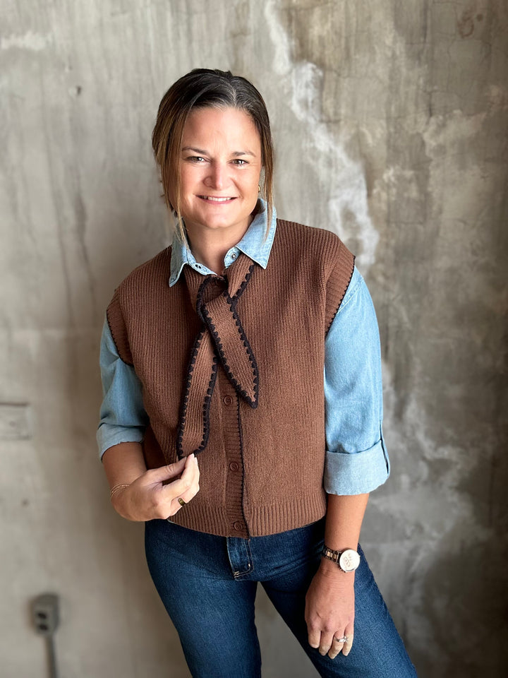 A woman wearing a brown knit button-front vest with black trim over a light blue collared shirt and blue jeans, standing against a textured concrete wall.