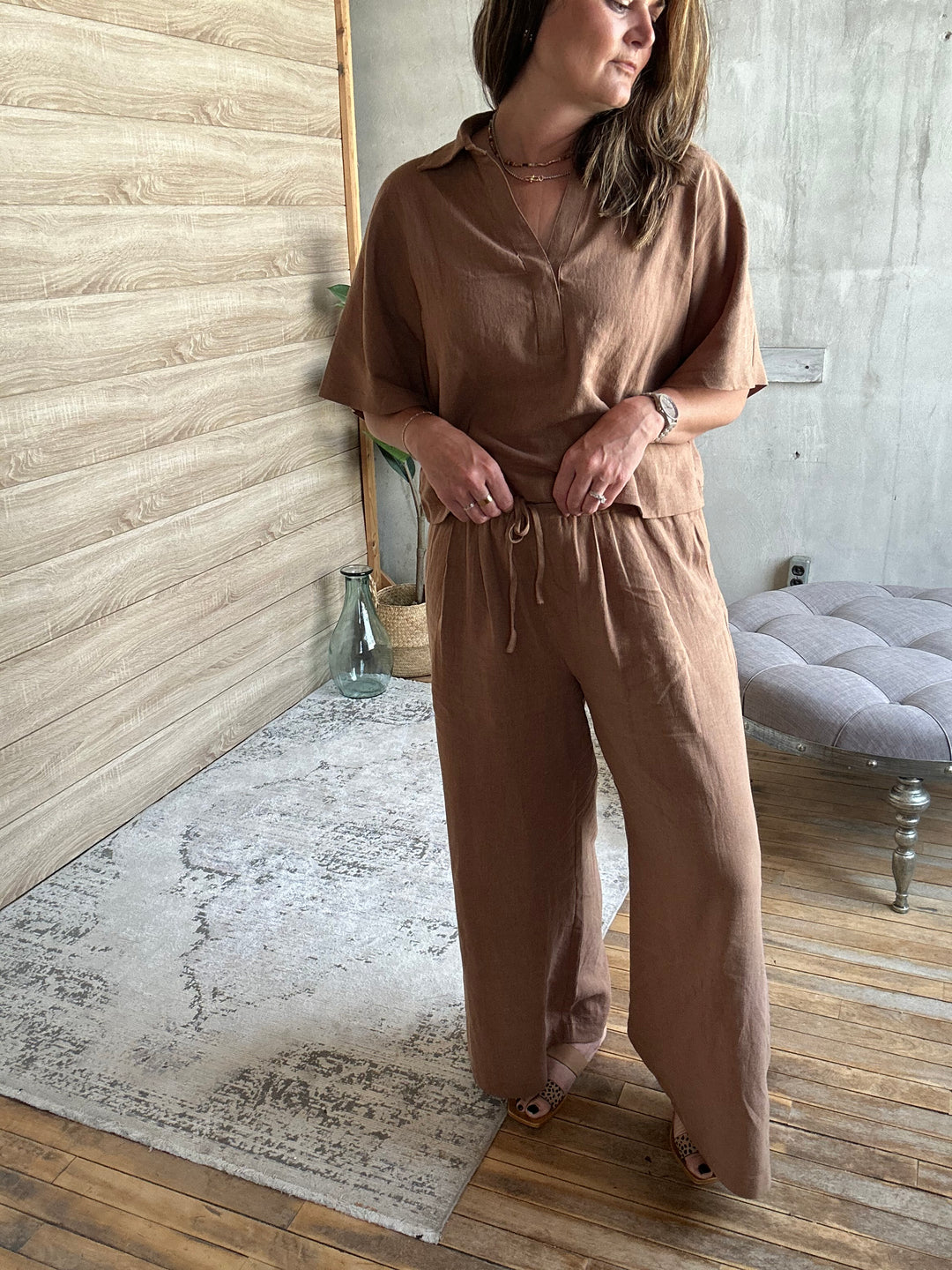 Woman in a brown outfit standing in a room with wooden flooring and a gray upholstered chair.