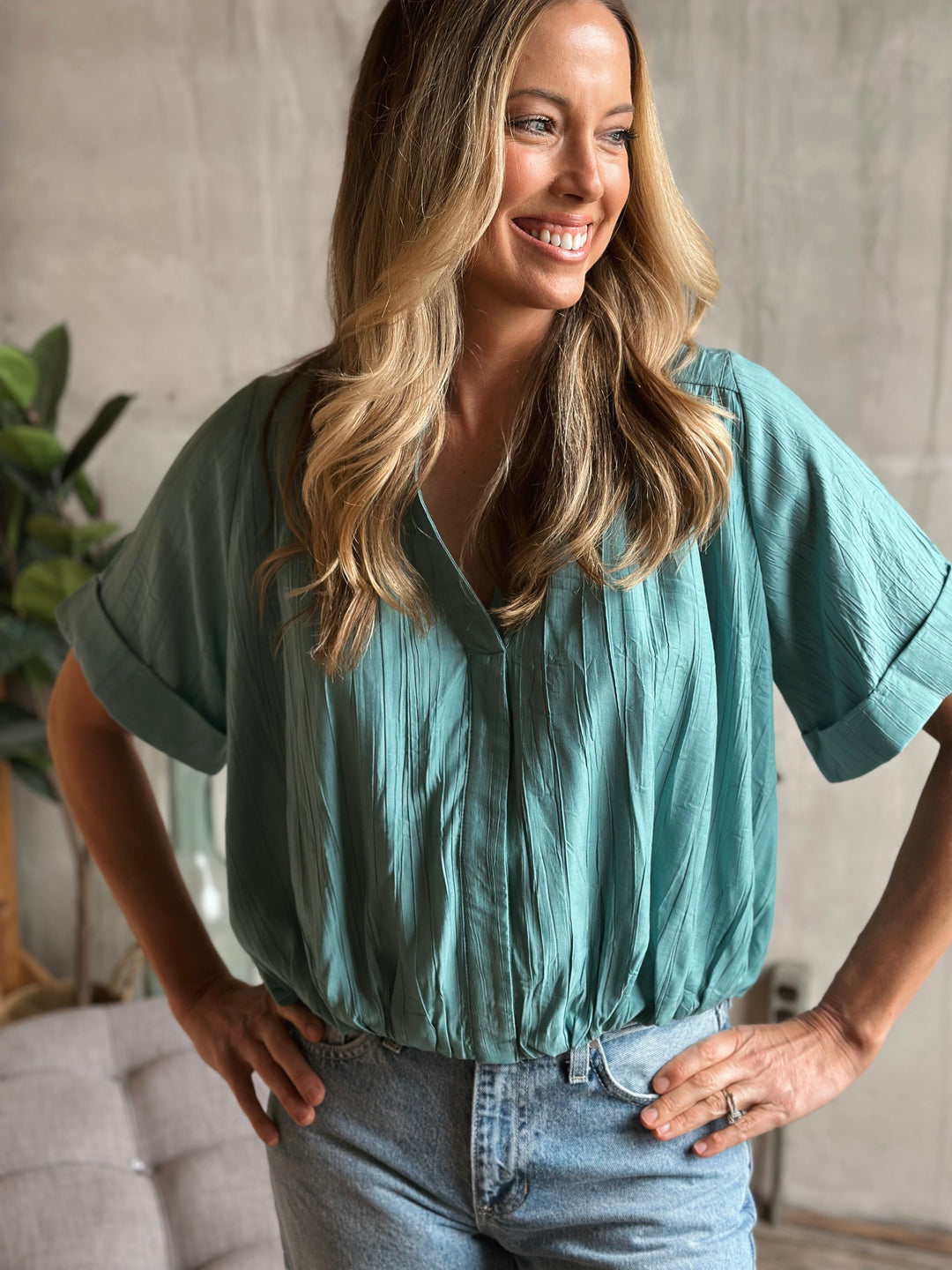 Woman wearing a teal blouse and blue jeans standing indoors with a neutral background.