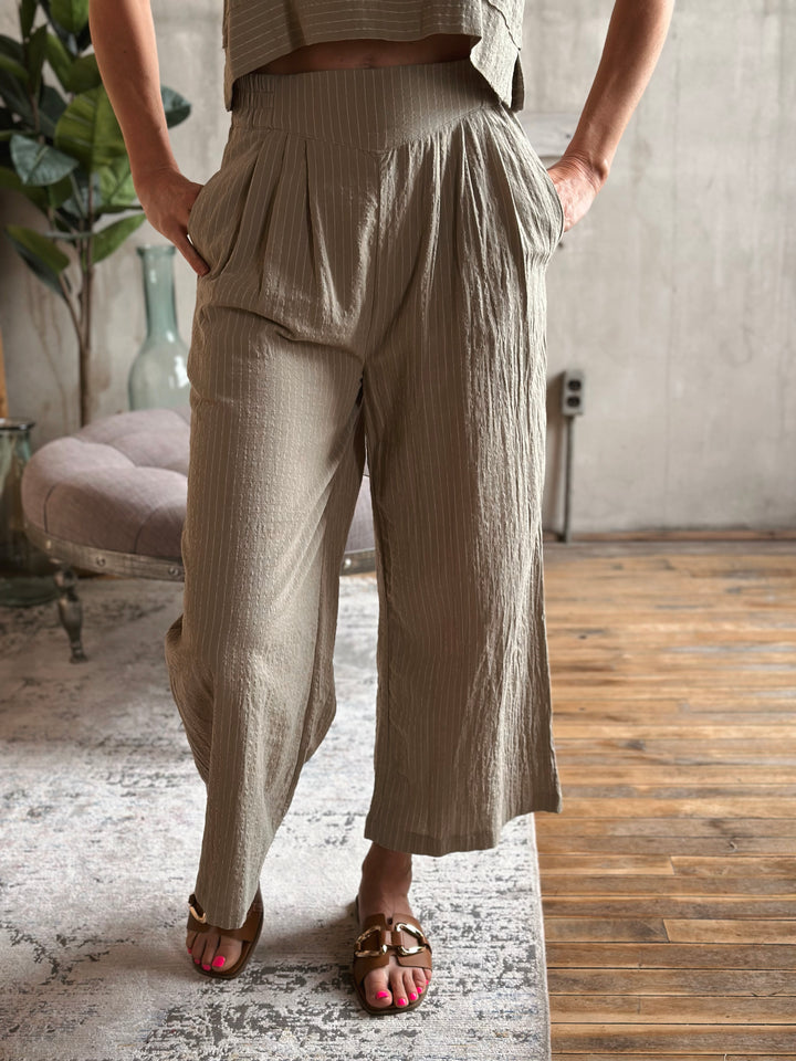 A person wearing sage wide-leg pants in a room with a wooden floor and plant.