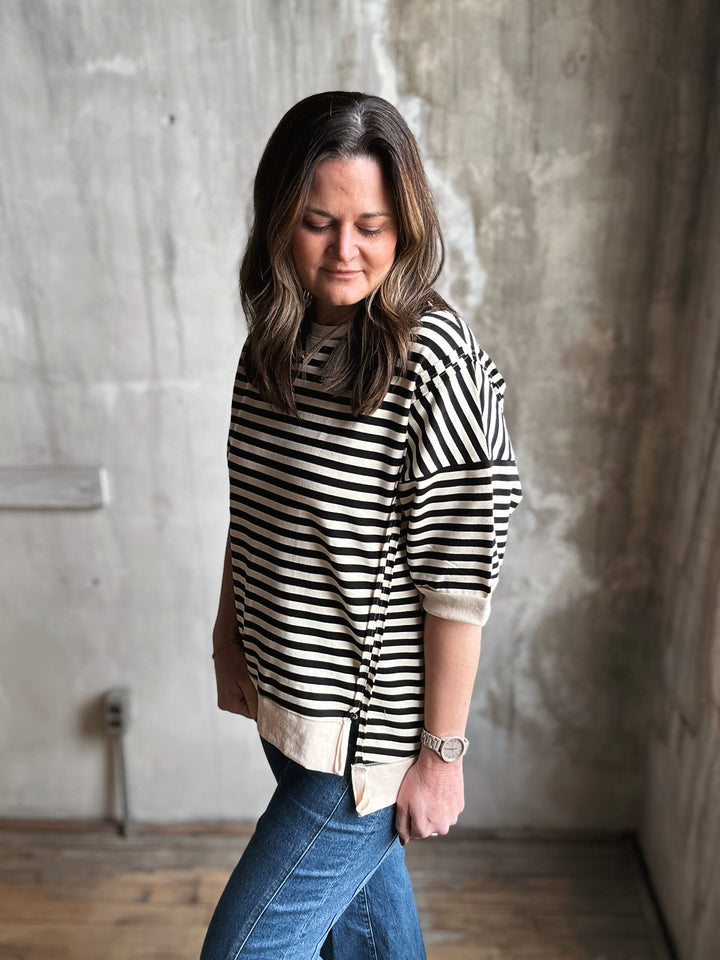Woman wearing a black and white striped sweater and blue jeans against a concrete wall.