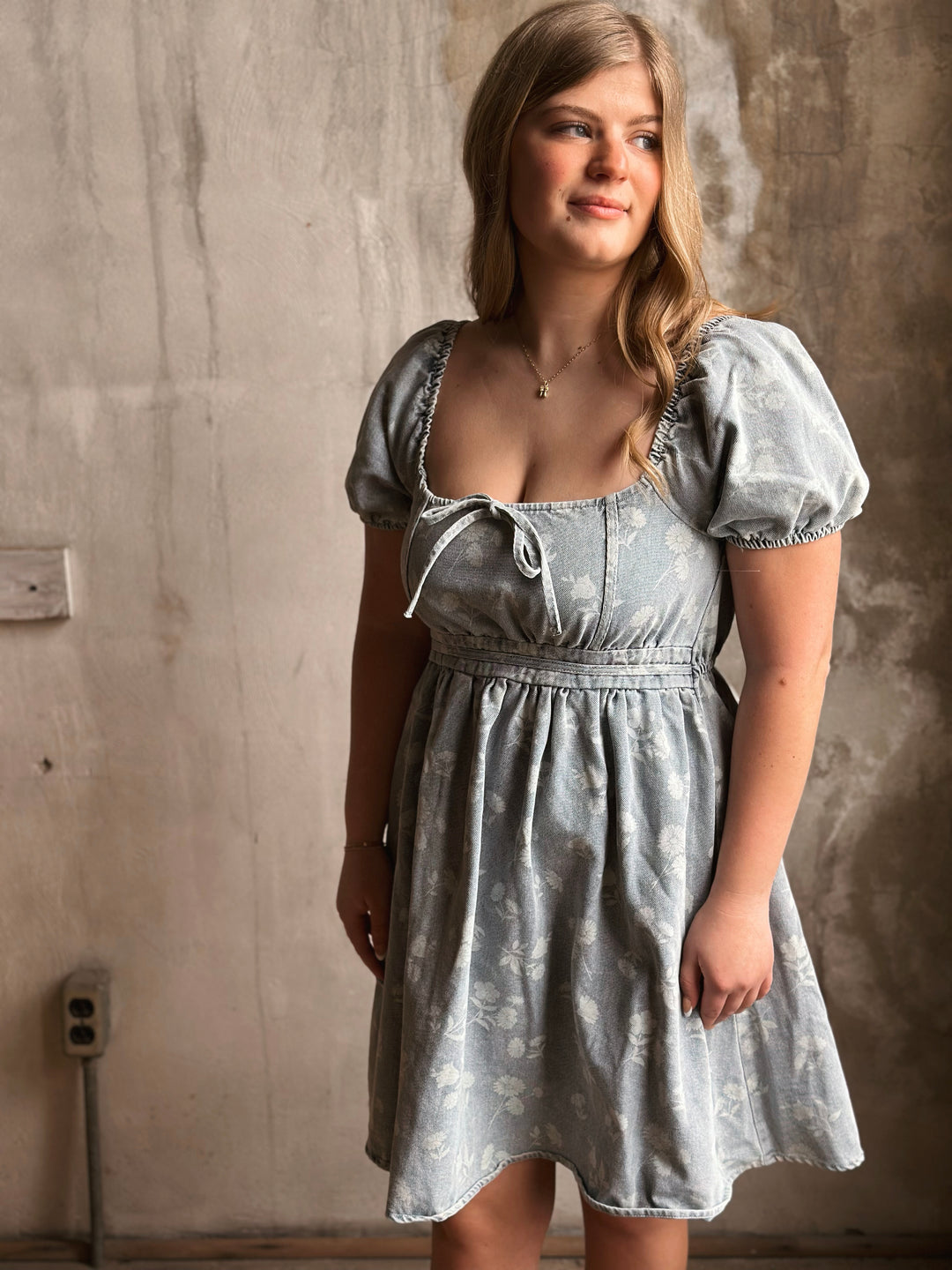 Woman wearing a light blue denim dress against a textured wall.