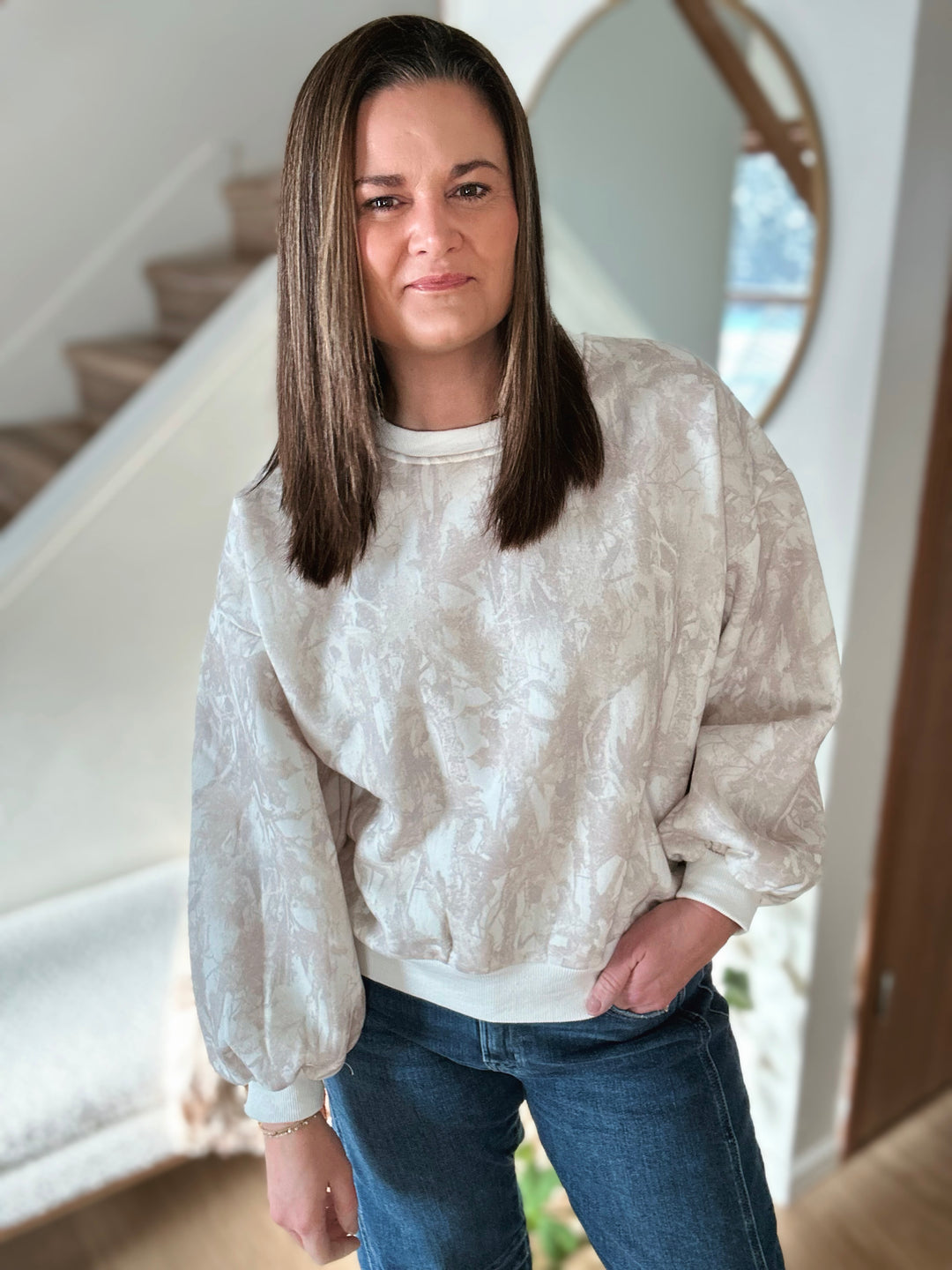 Woman wearing a white woodland sweatshirt and blue jeans standing in a home setting.
