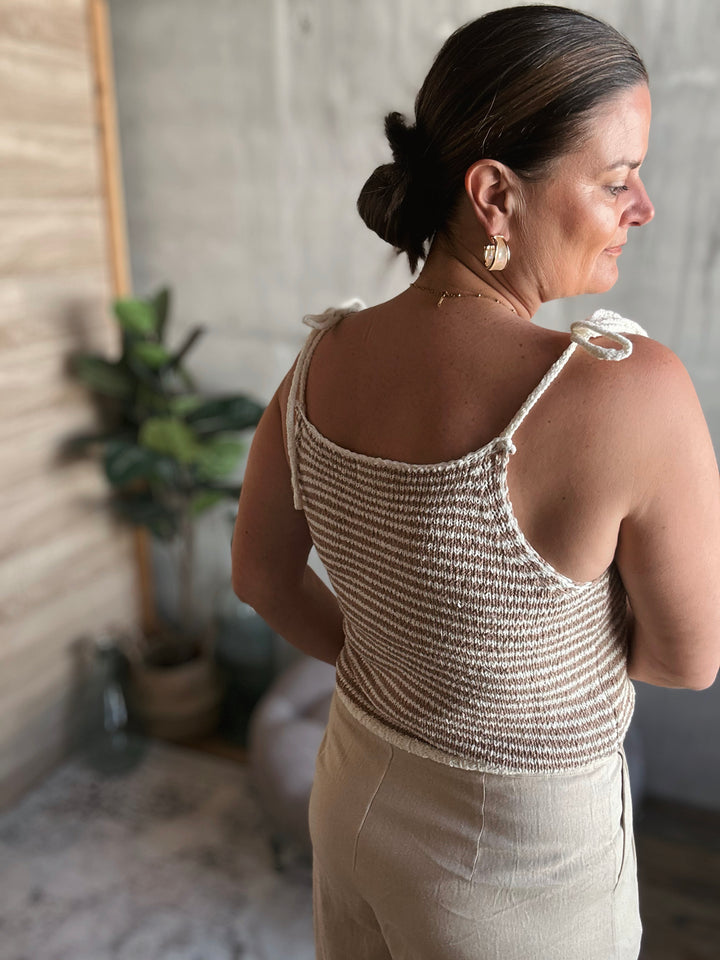 A woman wearing a light brown and beige striped crocheted tank top with thin straps that are tied into small bows.