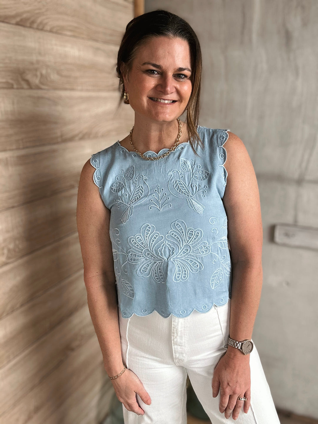 Woman wearing a light blue embroidered top and white pants against a neutral wall.