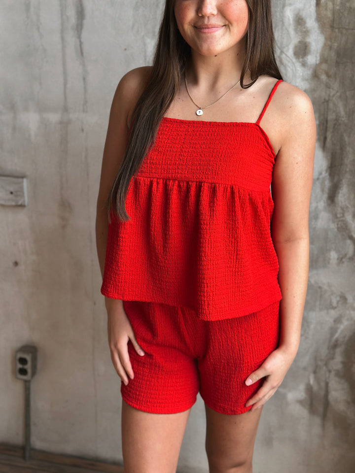 A person wearing a red top with thin straps  and shorts set against a textured wall.
