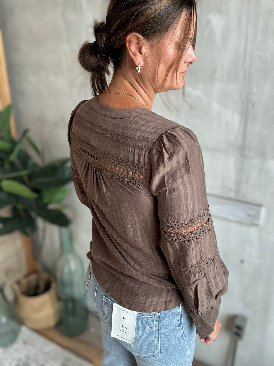 A woman wearing a brown long-sleeve blouse with lace-up back detailing and gathered sleeves, paired with light blue jeans, shown from a side-back angle in an indoor setting.