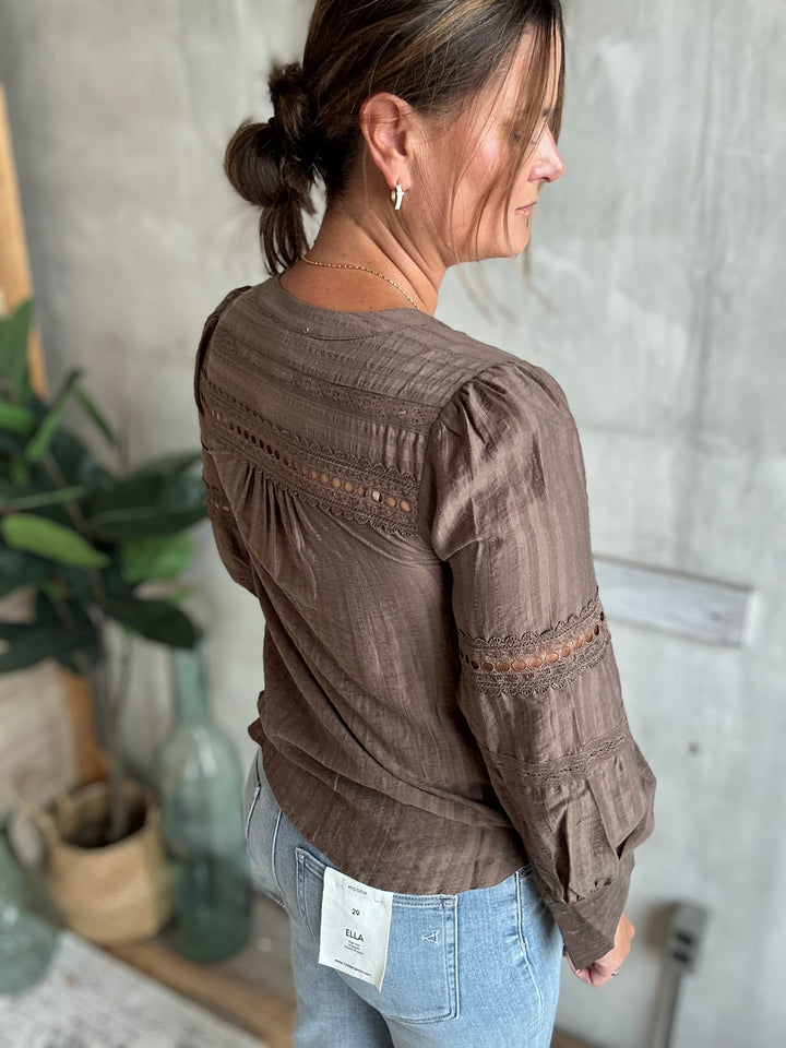 A woman wearing a brown long-sleeve blouse with lace-up back detailing and gathered sleeves, paired with light blue jeans, shown from a side-back angle in an indoor setting.