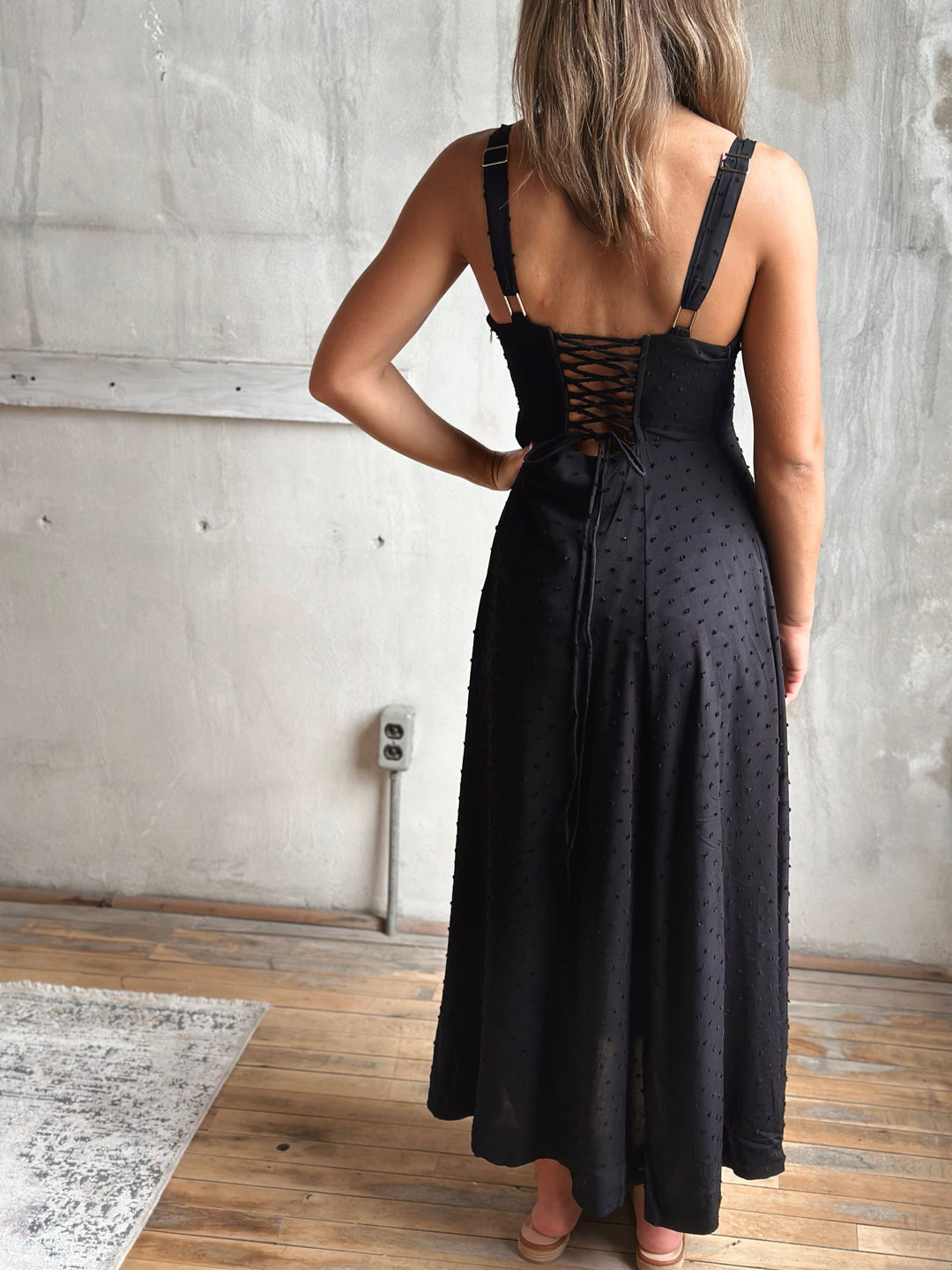 Person wearing a black dress with lace-up back in a room with concrete wall and wooden floor.