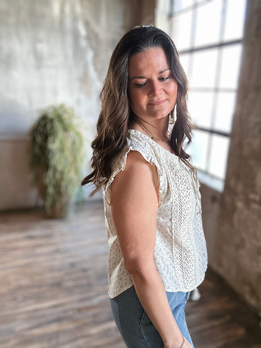 Side view of model wearing floral printed lace trim blouse with ruffle details and a front bow tie, it’s a sleeveless design with a round neckline and lace trim accents, this blouse is cream with light blue floral print