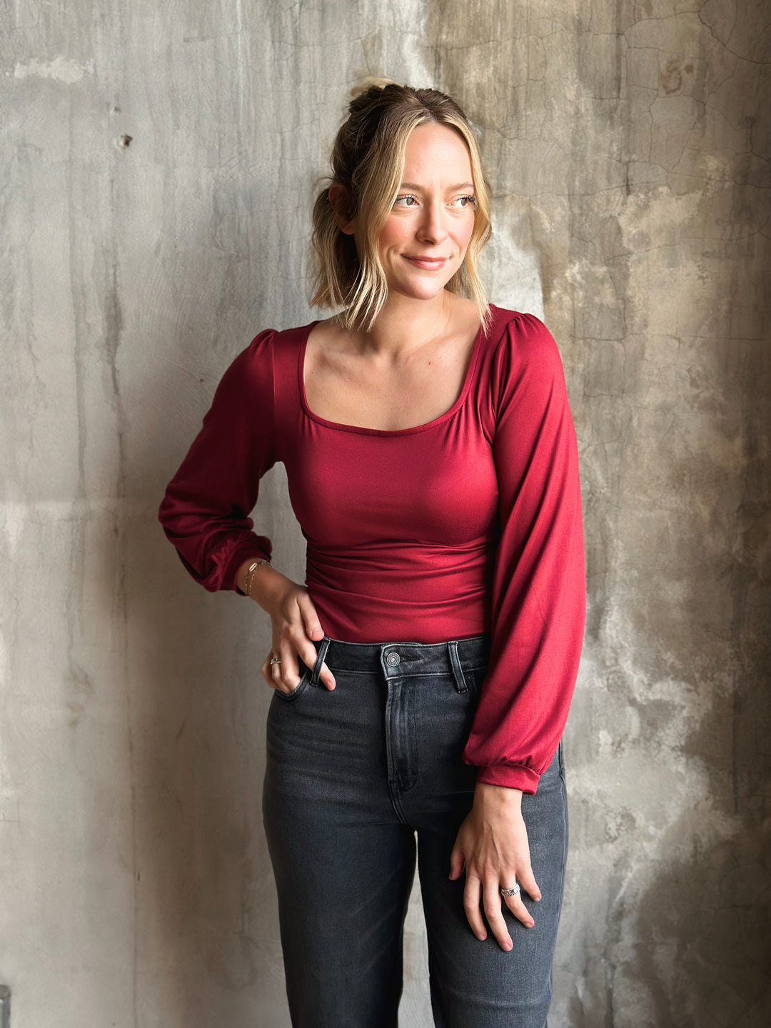 Woman wearing a red long-sleeve top and dark jeans against a textured wall.