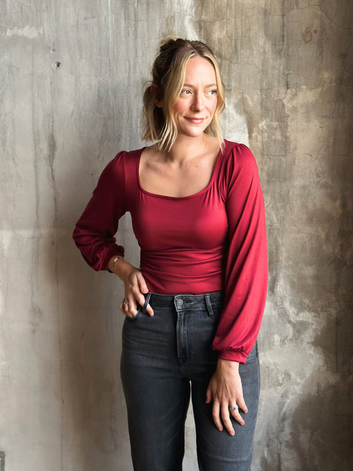 Woman wearing a red long-sleeve top and dark jeans against a textured wall.