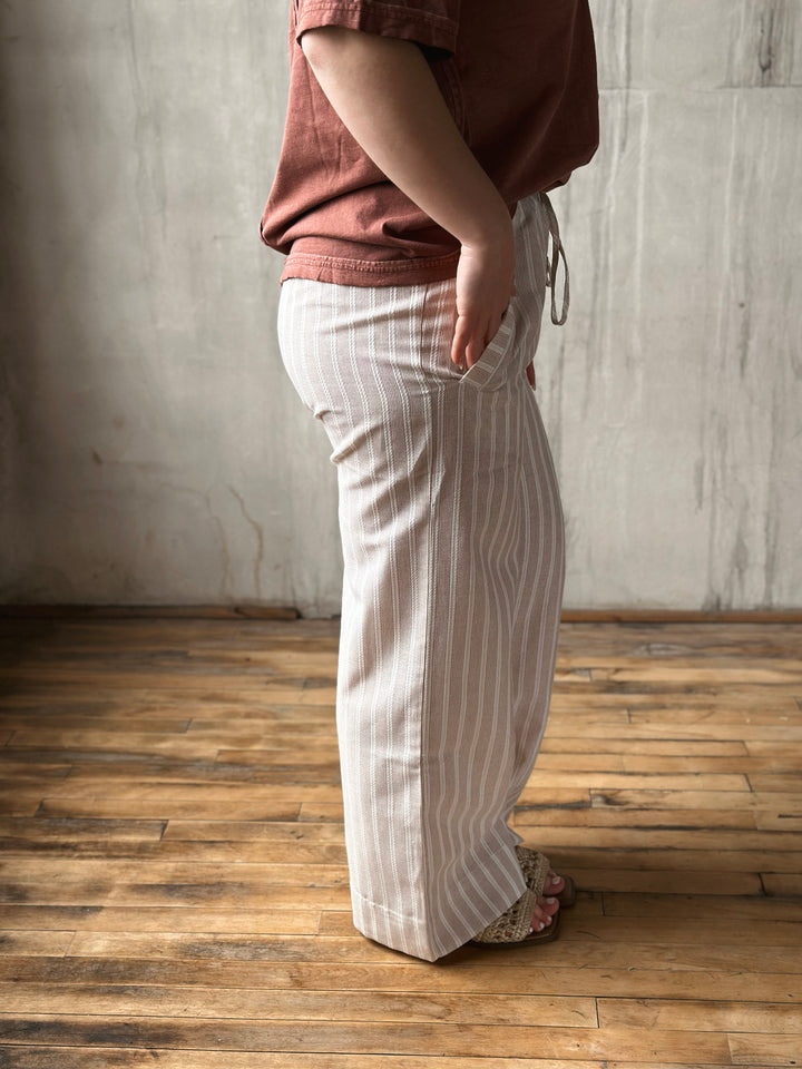 Person wearing a brown shirt and white striped pants standing on a wooden floor with a gray wall background.