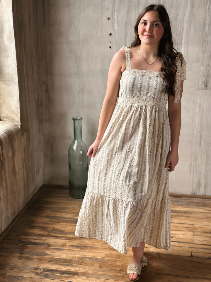Woman in a white lace dress with floral designs standing in a room with wooden floor and concrete wall.