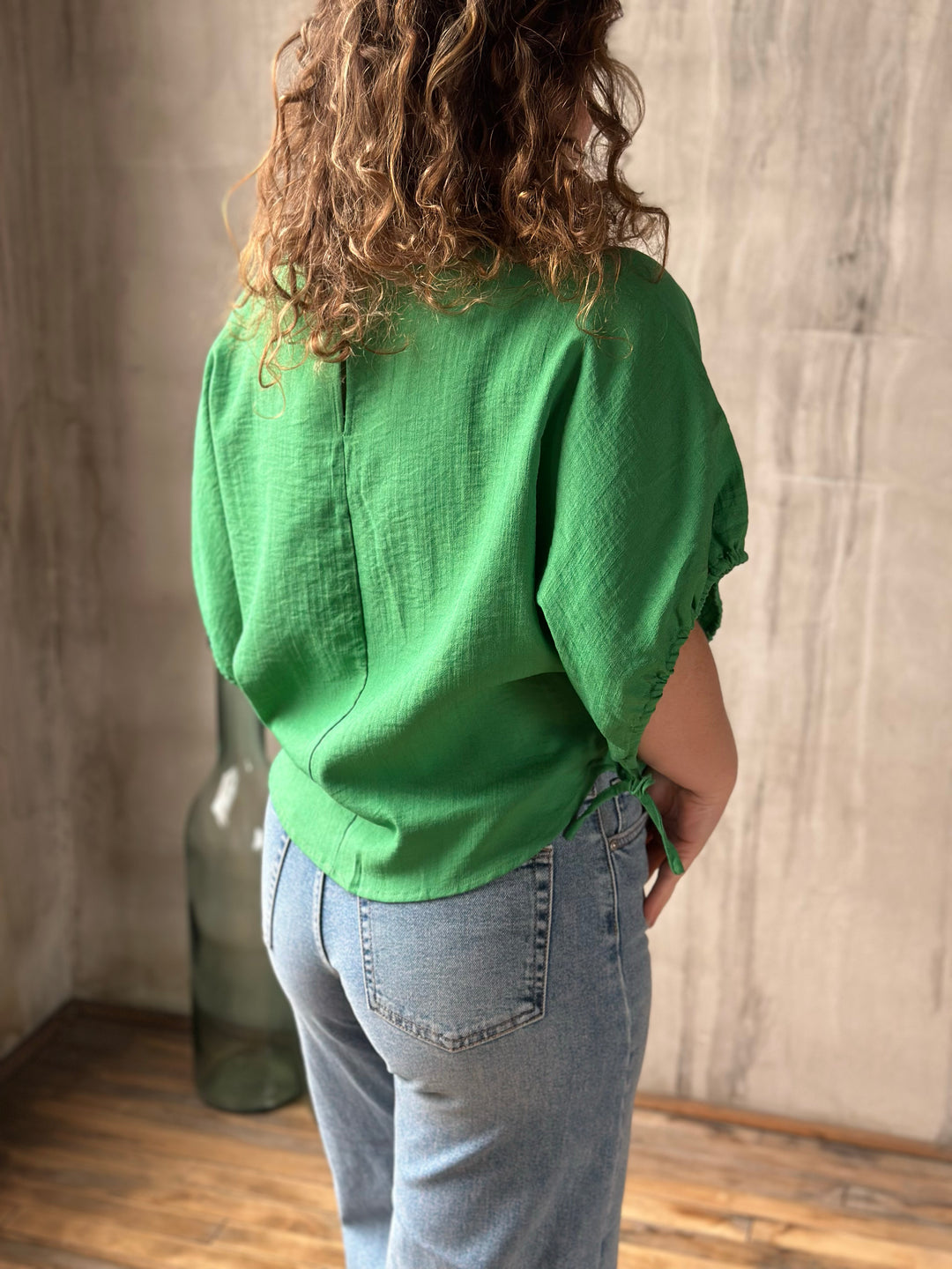 Person wearing a green blouse and blue jeans standing against a wooden floor and wall.