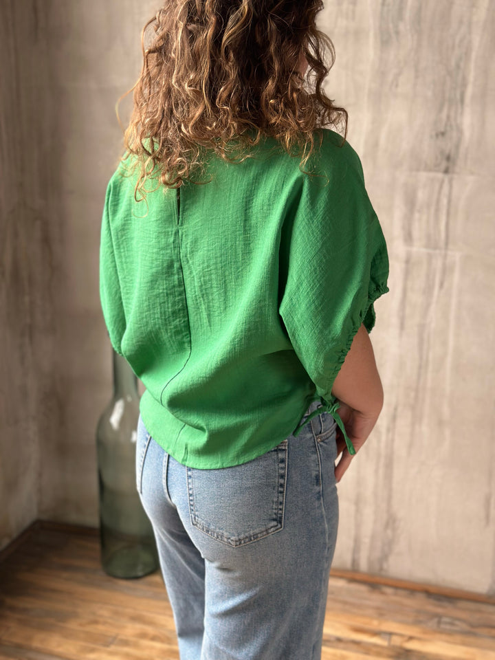 Person wearing a green blouse and blue jeans standing against a wooden floor and wall.