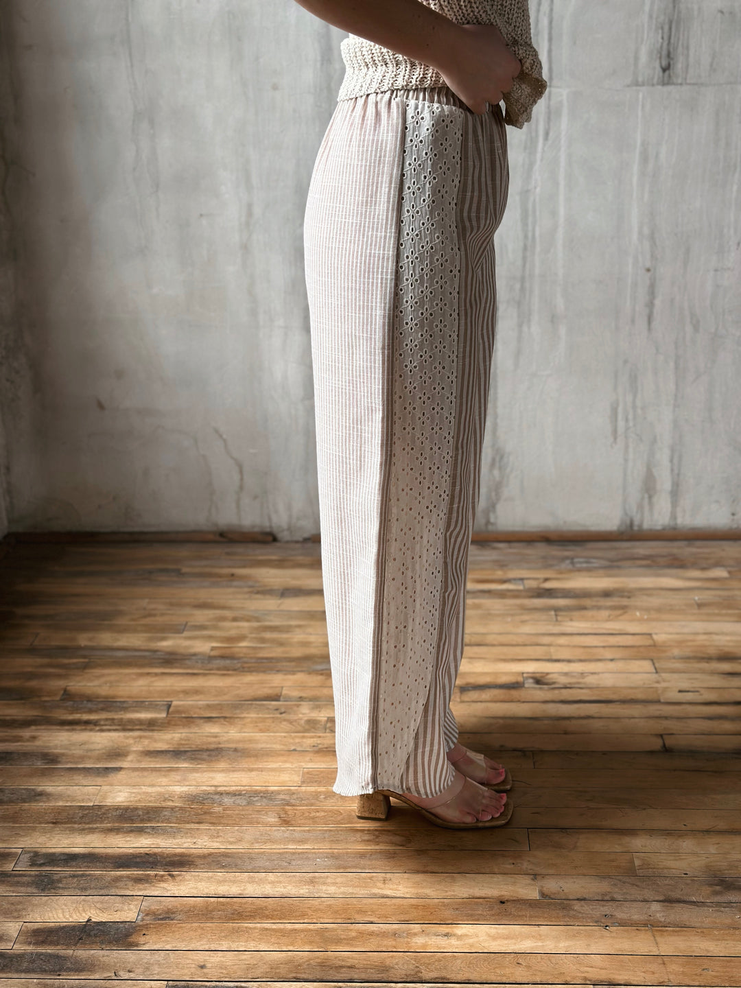 Person wearing striped pants with a lace detail on the side, standing on a wooden floor with a gray wall background