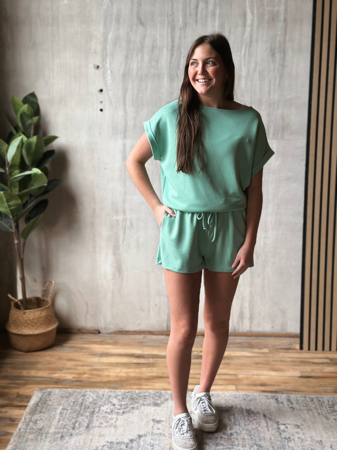 Woman wearing a green outfit standing in a room with a plant and concrete wall.