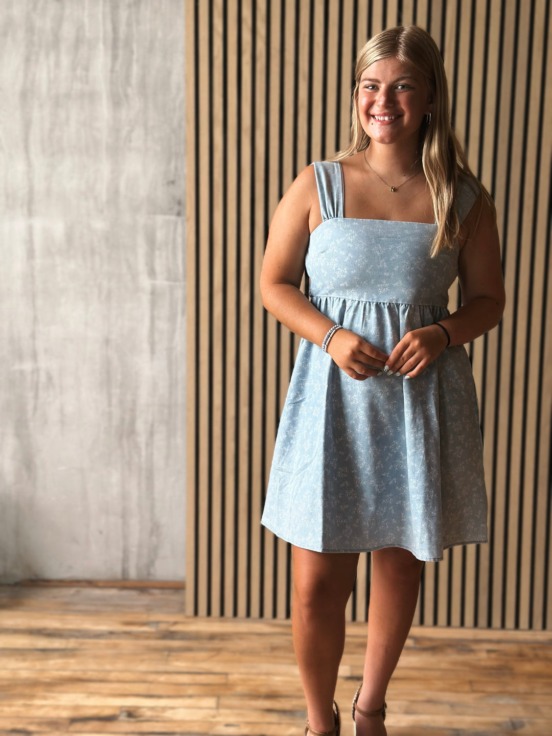 Woman wearing a light blue dress standing against a wooden paneled wall.