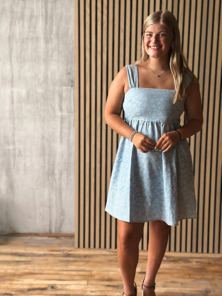 Woman wearing a light blue dress standing against a wooden paneled wall.