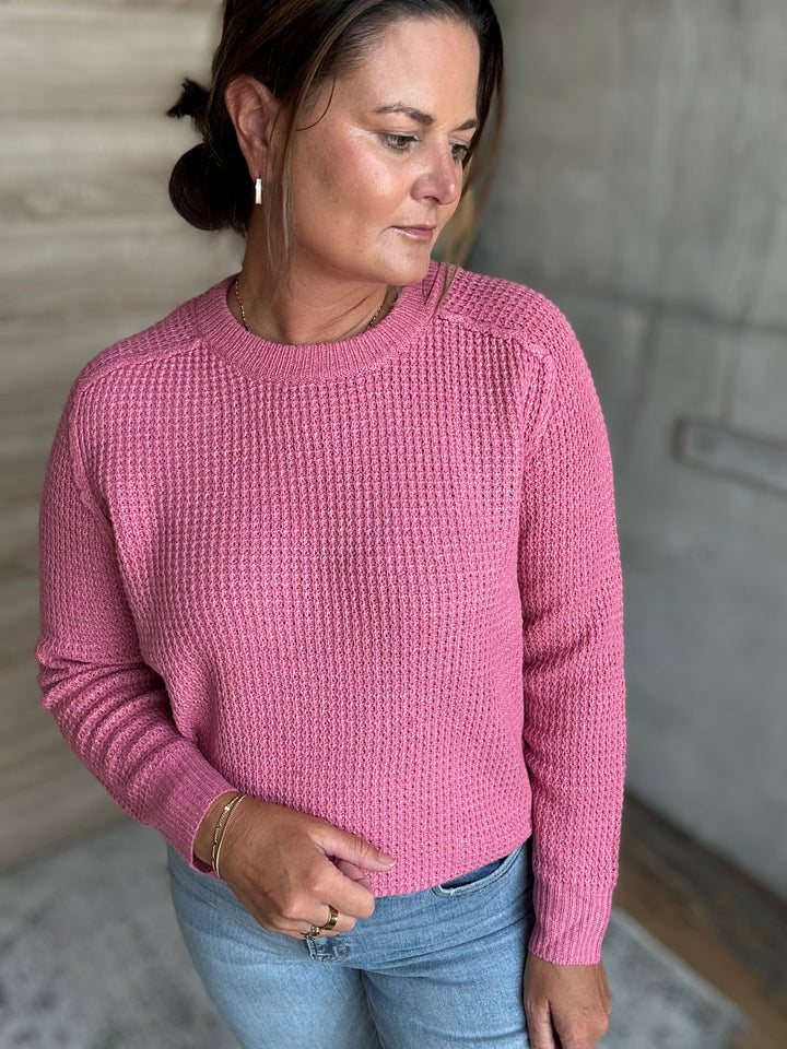 A woman wearing a textured waffle-knit long-sleeve sweater in dusty pink, paired with light blue jeans, standing against a neutral gray concrete wall.