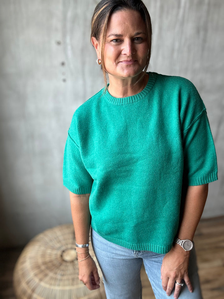 A woman wearing a vibrant green short-sleeve knit sweater and light blue jeans, standing in front of a textured concrete wall.