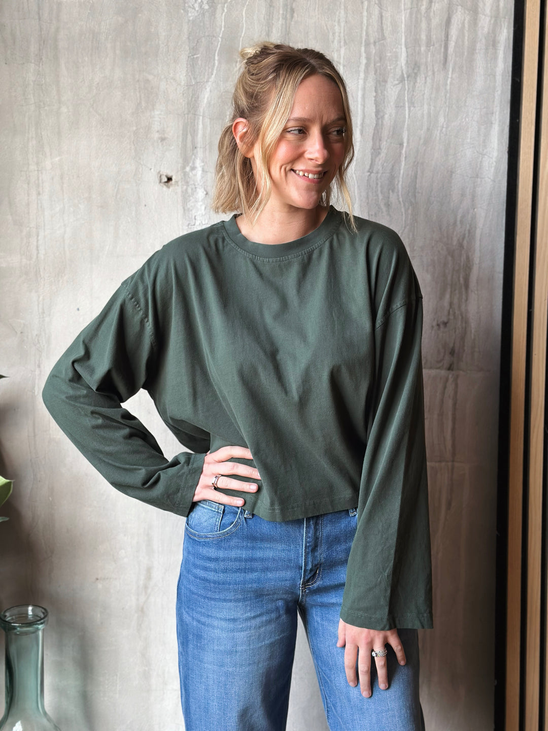 Woman wearing a green long-sleeve top and blue jeans against a wooden wall.