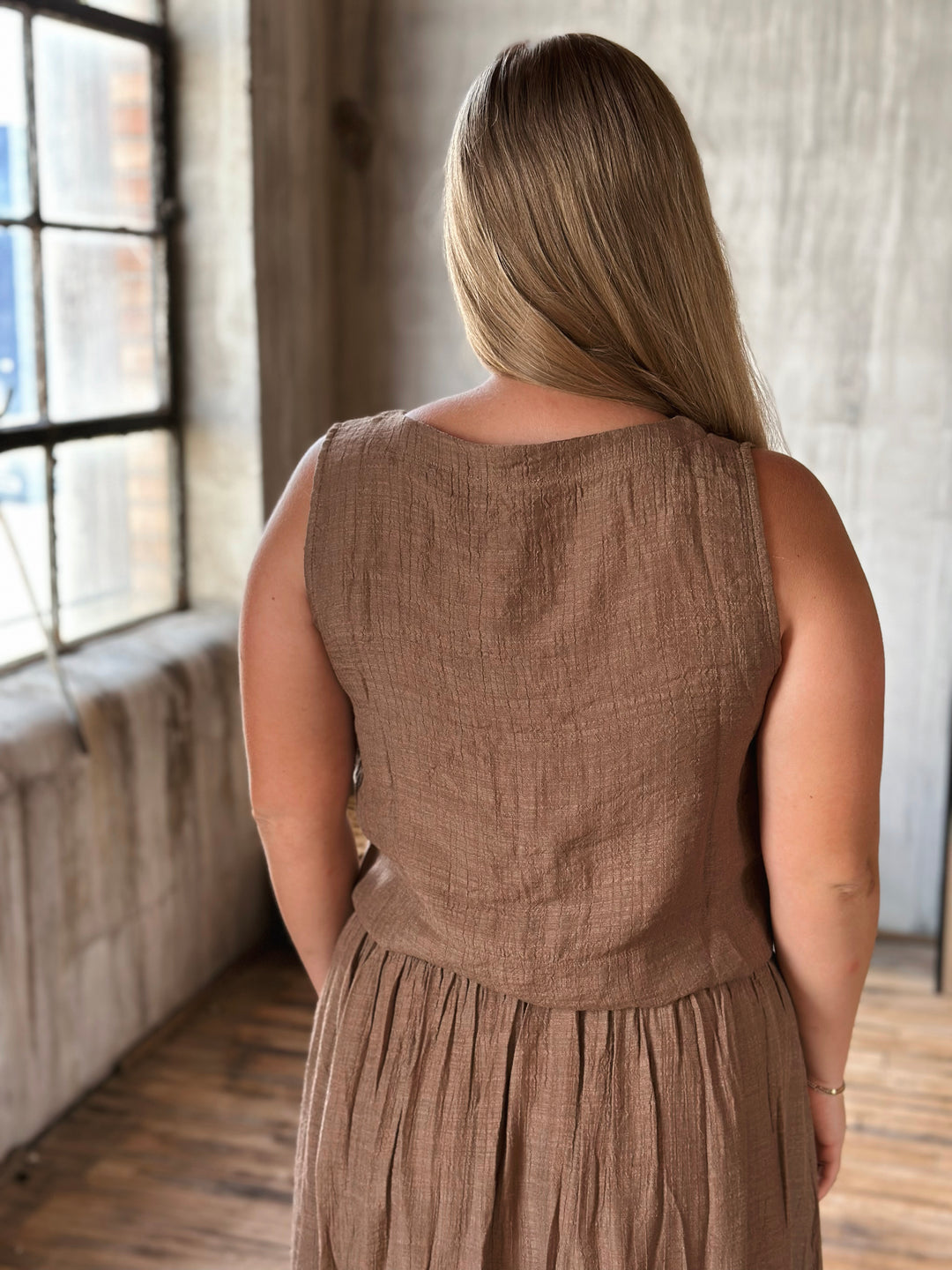 Woman wearing a brown sleeveless top standing in a room with large windows.