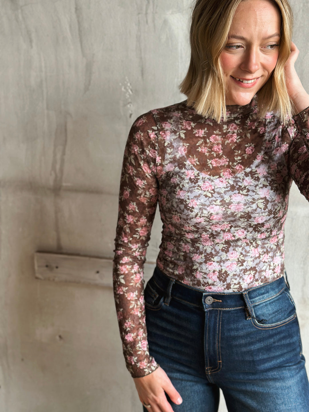 Woman wearing a floral long-sleeve top and jeans against a concrete wall.