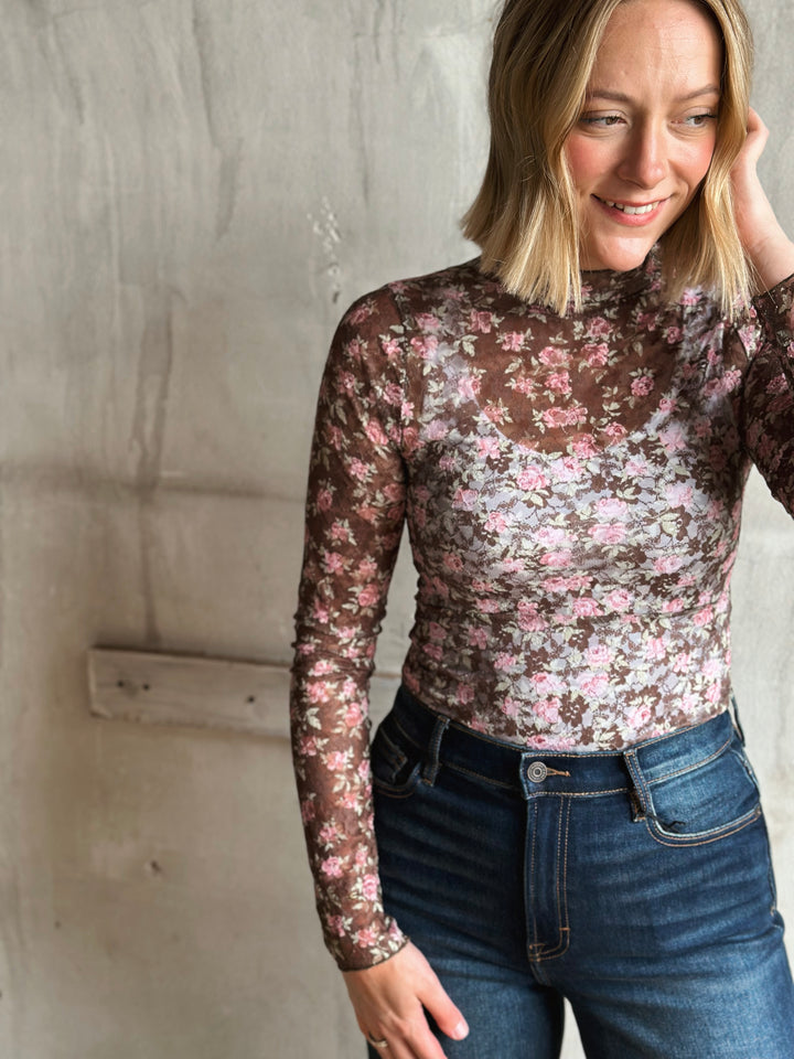 Woman wearing a floral long-sleeve top and jeans against a concrete wall.