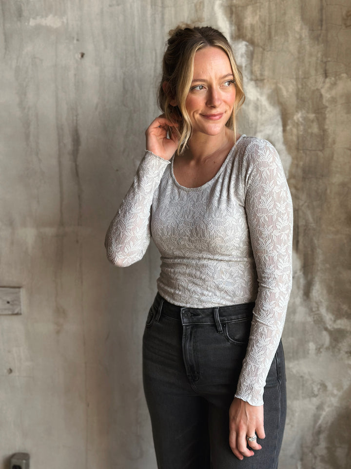 Woman wearing a light gray long-sleeve top and dark jeans against a textured wall.