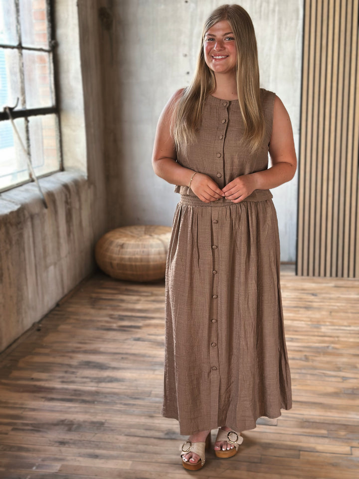 A woman wearing a brown skirt with buttons and a matching top standing in a room with a wooden floor and large window.