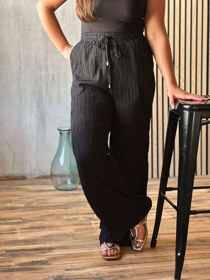 Person wearing black wide-leg pants standing next to a black stool indoors.